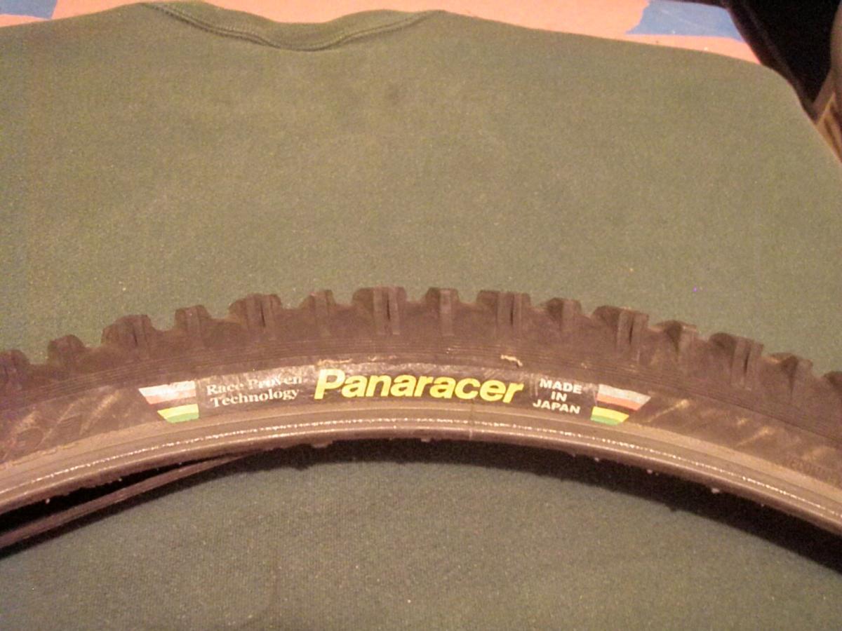 Panaracer Smoke: A close-up view of a mountain bike tire featuring the brand name "Panaracer," with text indicating "Made in Japan" and mentions of "Race Trusted Technology." The tire shows a rugged tread pattern, suitable for off-road use, and is placed on a green fabric background.