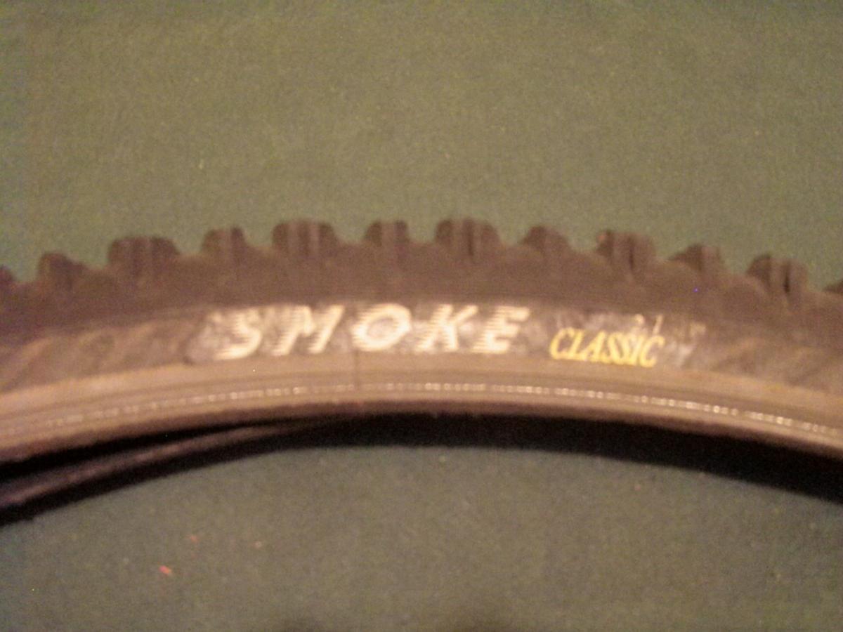 Panaracer Smoke: A close-up view of a bicycle tire showcasing the tread pattern and branding, with the words "SMOKE" and "CLASSIC" visible on the sidewall. The tire is positioned on a green surface.