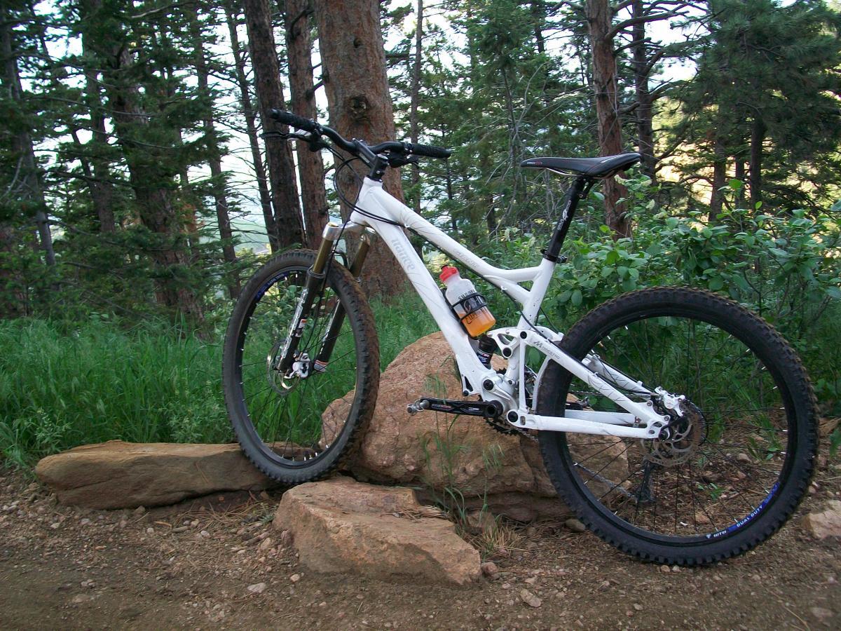 Giant Trance 1: A white mountain bike positioned on a rocky surface, surrounded by tall trees and lush greenery, on a dirt trail in a forest setting. The bike has a water bottle attached to the frame and is angled to showcase its features against the natural backdrop.
