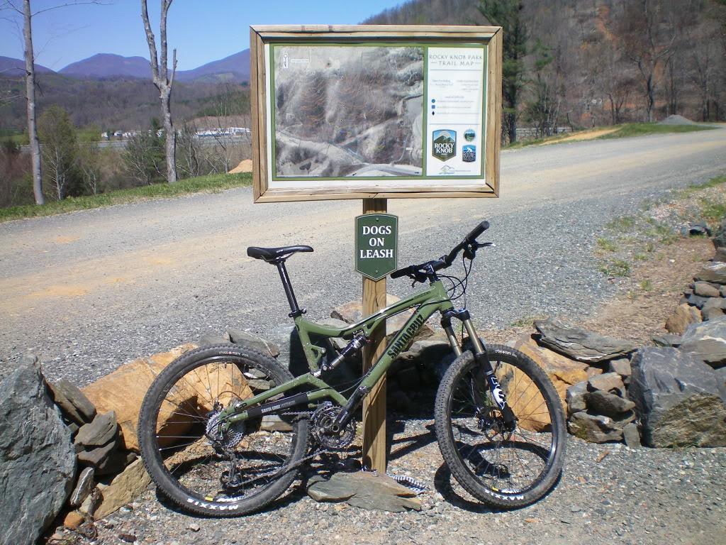 Santa Cruz Nickel: A mountain bike parked next to a trail information sign that reads "Dogs on Leash." The scene includes a gravel road, a landscaped area with stone features, and distant mountains under a clear blue sky.