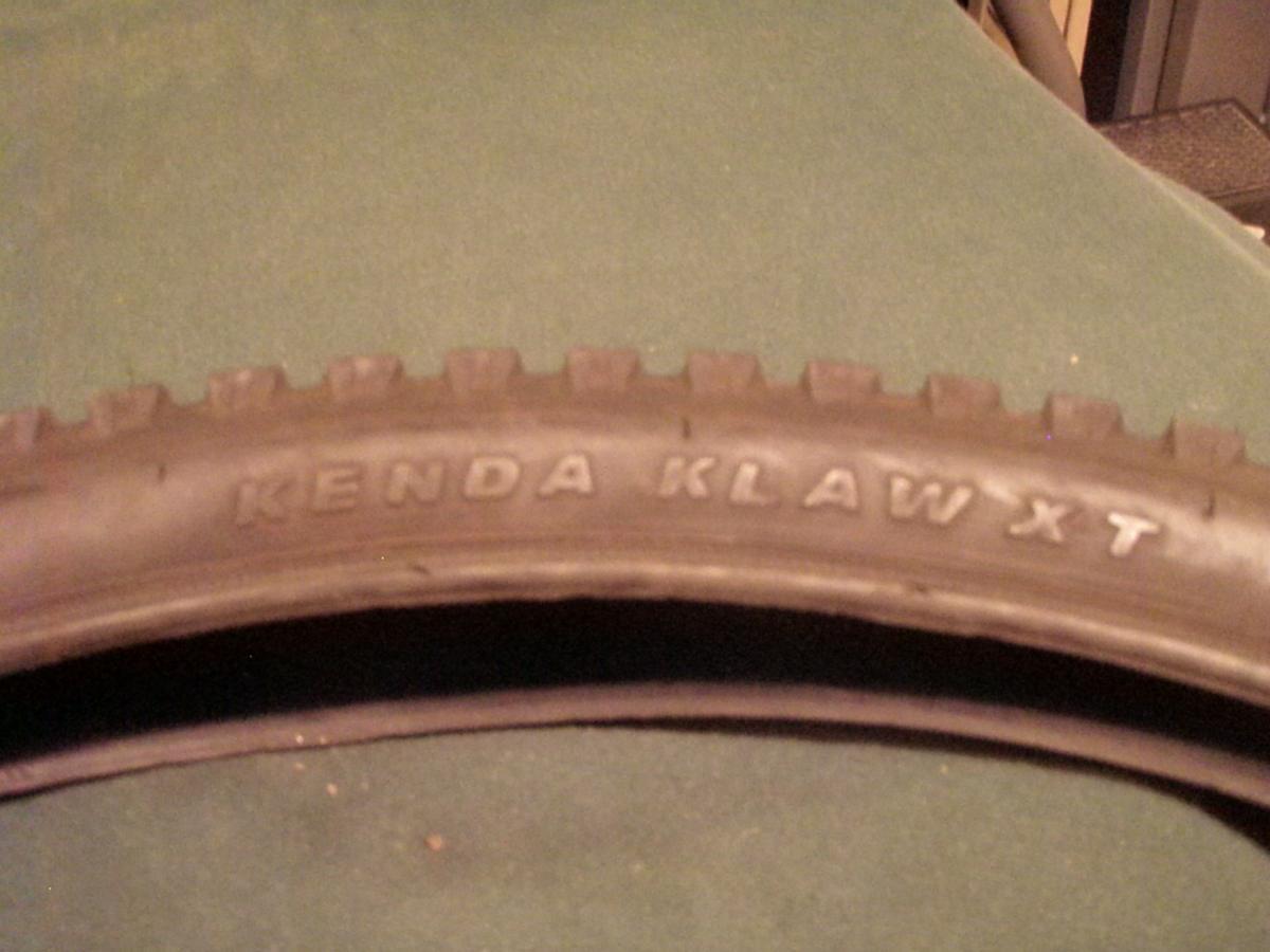 Kenda Klaw: Image of a Kenda Klaw XT bicycle tire resting on a green surface, showcasing the tire tread and brand name embossed on the sidewall.