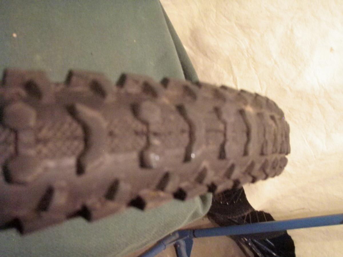 Kenda Klaw: Close-up image of a textured tire, showcasing its tread pattern and details. The tire is positioned on a green surface, with some soft shadows visible.