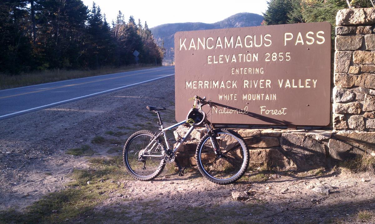 Gary Fisher Marlin: A mountain bike leaning against a stone sign that reads "Kancamagus Pass, Elevation 2855, Entering Merrimack River Valley, White Mountain National Forest," with a road and forested mountains in the background.