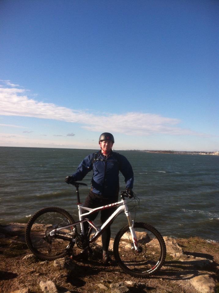 A person wearing a helmet stands next to a mountain bike on rocky terrain by a body of water. The sky is clear with a few clouds, and the scene captures a sense of outdoor adventure. Bluff Point State Park mountain bike trail.