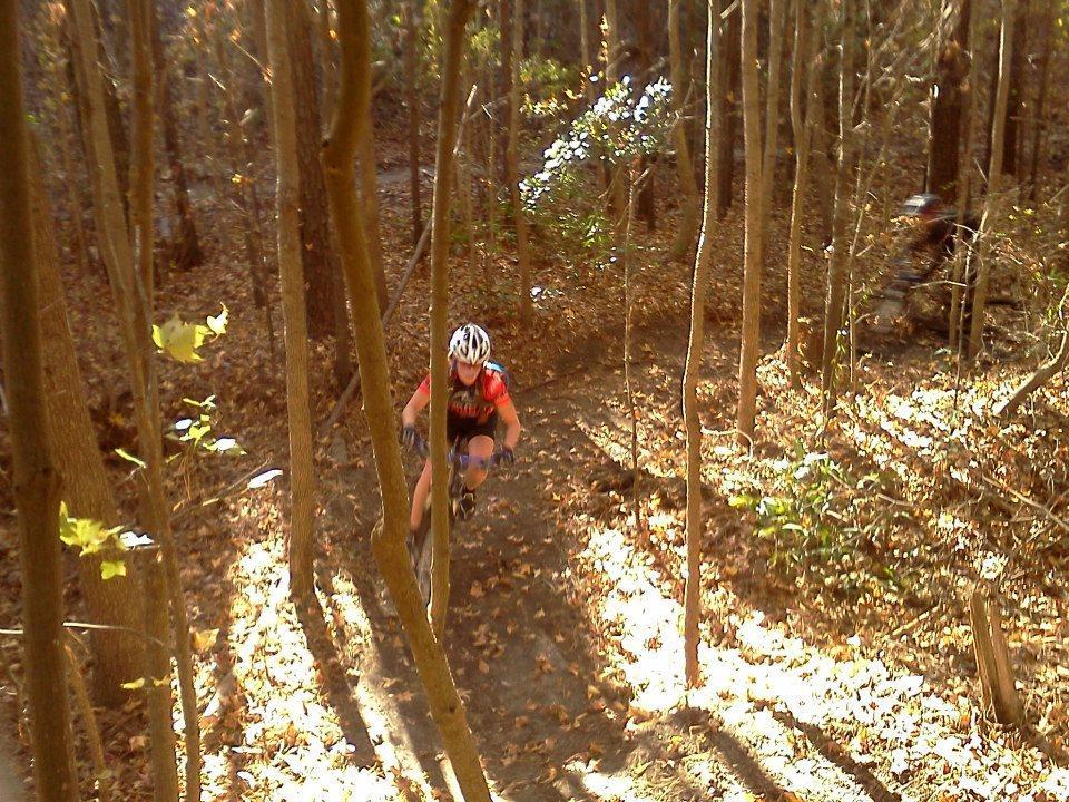 A mountain biker navigating a dirt trail through a forest with autumn foliage. Sunlight filters through the trees, illuminating the scene. The biker is wearing a helmet and cycling gear, focused on the ride. Horry County Bike Run Park mountain bike trail.