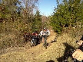 Two mountain bikers navigate a dirt trail surrounded by greenery and trees. The scene captures a moment of outdoor adventure, with one rider climbing a slope while the other follows closely behind. Bright sunlight filters through the trees, illuminating the natural setting. Horry County Bike Run Park mountain bike trail.