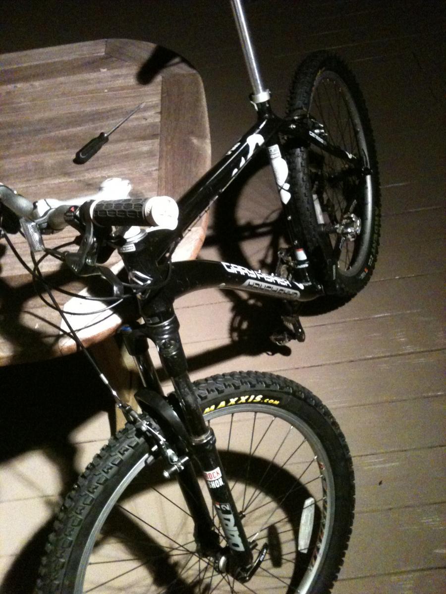Gary Fisher Marlin: A close-up image of a mountain bike in low light conditions, featuring a black frame with white accents, prominent tires suitable for off-road use, and various components like handlebars and a fork. A screwdriver is resting on a wooden surface nearby.