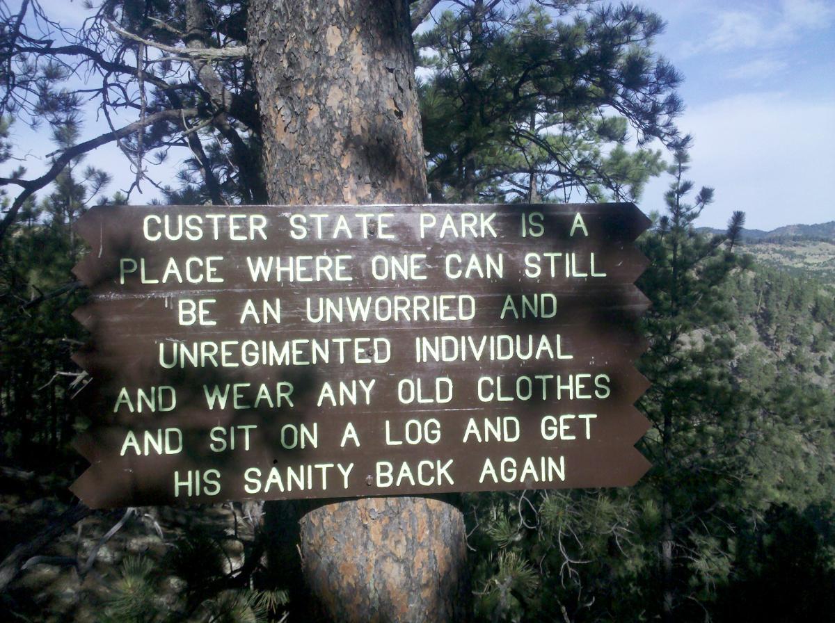 A wooden sign at Custer State Park with text that reads: "Custer State Park is a place where one can still be an unworried and unregimented individual and wear any old clothes and sit on a log and get his sanity back again." The sign is mounted on a tree with greenery and hills visible in the background. Lovers Leap Trail mountain bike trail.