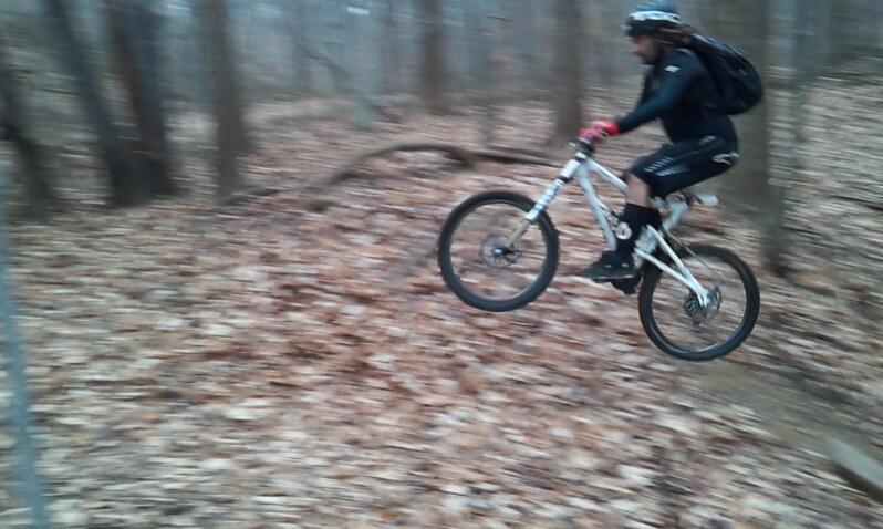 A person in cycling gear jumps off a small ramp on a mountain bike, surrounded by trees and fallen leaves in a forested area. The image is slightly blurred, conveying motion and excitement. Brandywine State Park mountain bike trail.