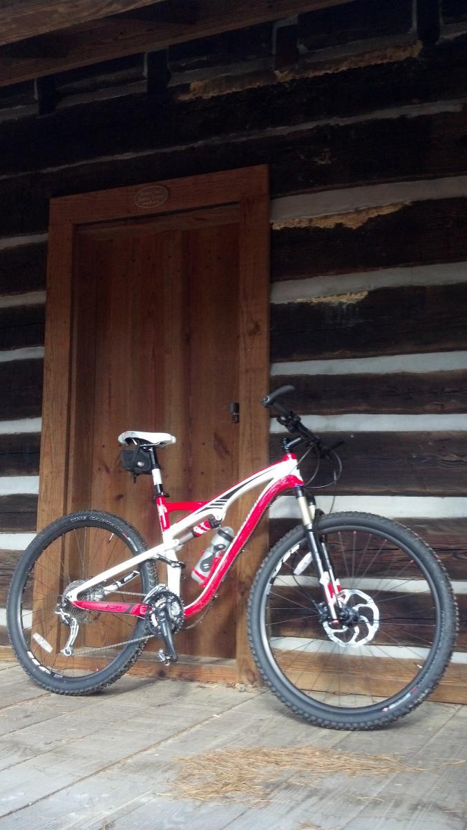 Specialized Camber 29: A mountain bike leaning against a wooden door of a rustic log cabin, with a wooden floor visible and some pine needles scattered around.