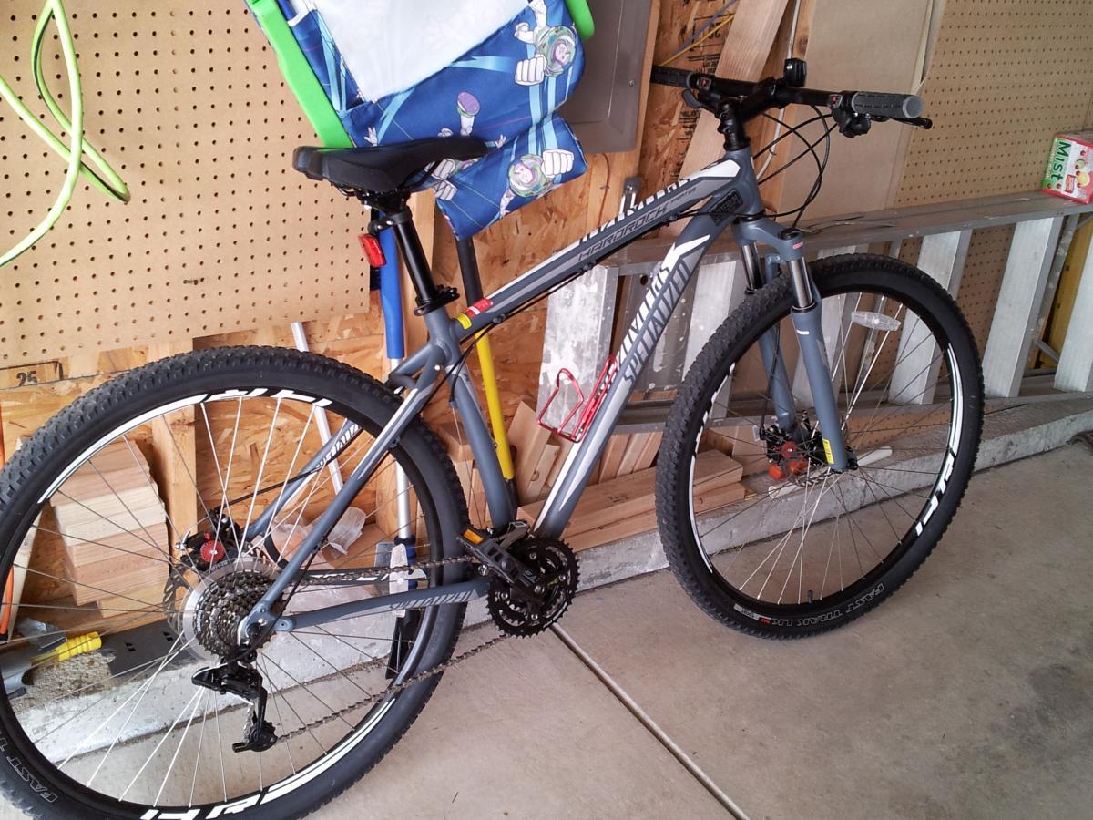Specialized Hardrock Sport Disc 29er: A gray mountain bike with a red and yellow bottle holder, resting on a concrete floor in a garage. Behind it, there are wooden boards and tools, with a colorful beach chair hanging on a pegboard. The bike features thick tires designed for off-road use.
