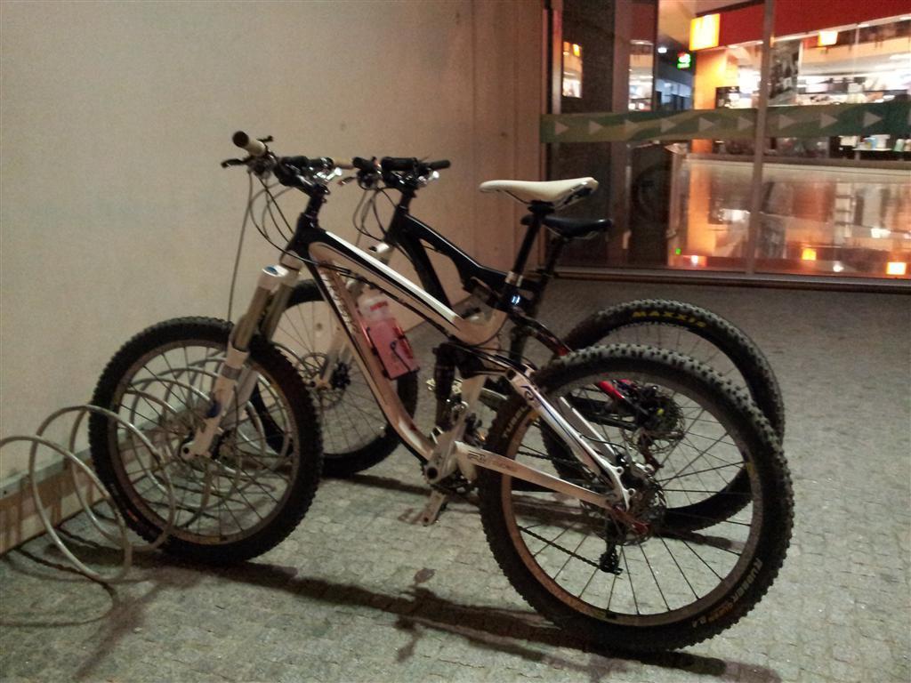 Trek Remedy 9: Two mountain bikes are parked next to a wall, with one bike mainly white and silver and the other black and white. The setting appears to be a well-lit area, possibly near a shopping or recreational space, with a glimpse of reflections from nearby windows.
