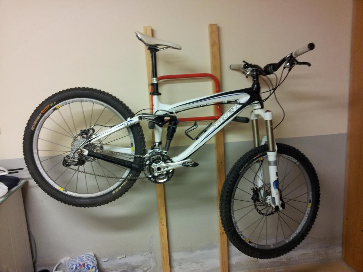 Trek Remedy 9: A white and black mountain bike hanging on a wall mount in a garage or storage area. The bike features thick tires, front suspension, and a visible gear system. The background shows a simple interior with a wooden support and a hint of other items on a shelf.