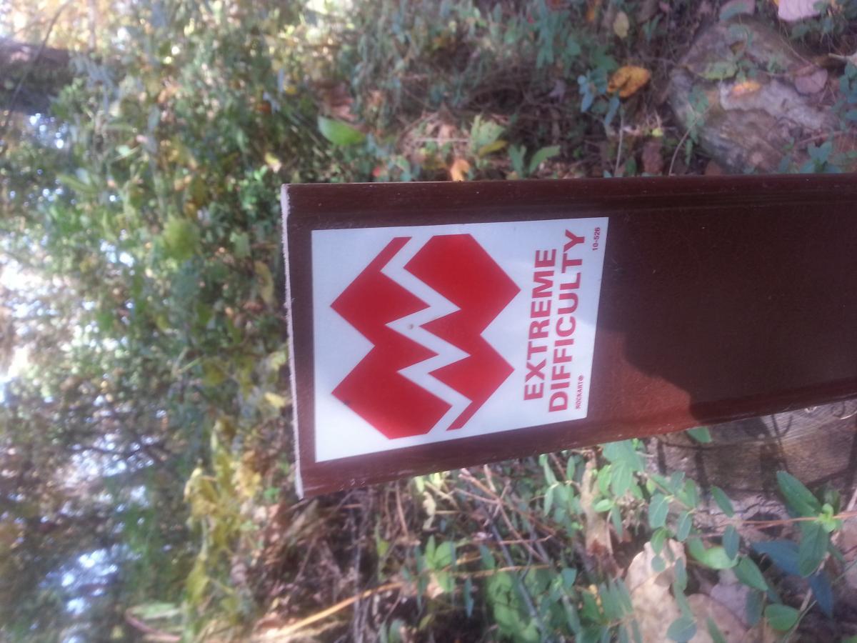 Specialized Epic Comp 29er: A brown trail sign displaying a red emblem with the words "EXTREME DIFFICULTY," set against a backdrop of greenery and forest foliage.
