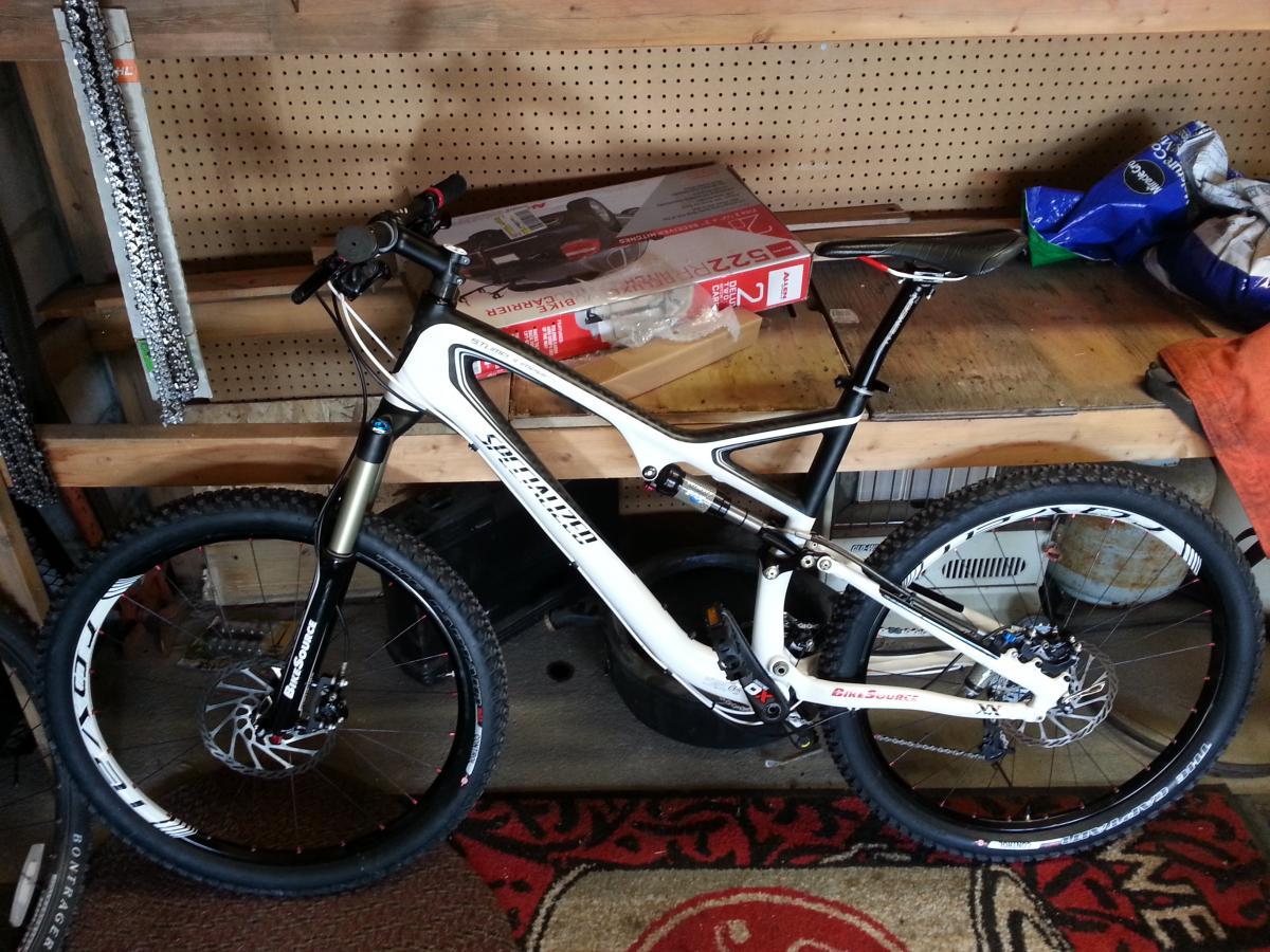 Specialized Stumpjumper FSR Pro Carbon: A mountain bike with a white and black frame is leaning against a wooden workbench in a garage. The bike features thick tires and has suspension forks in the front. In the background, various tools and items are visible on the workbench, including a cardboard box and some bags. The garage floor has a colorful rug.