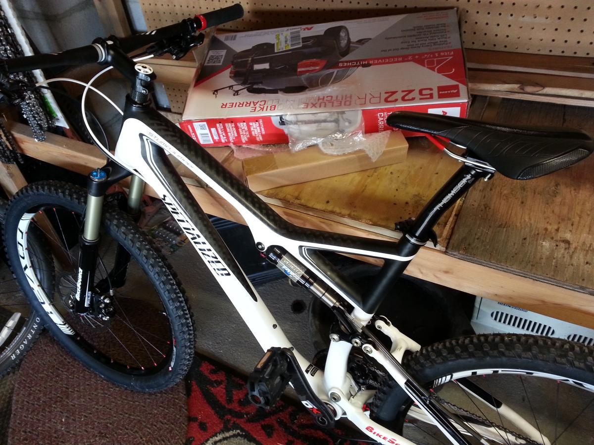 Specialized Stumpjumper FSR Pro Carbon: Image of a mountain bike resting on a workbench in a garage. The bike features a sleek white and black design, with thick tires suitable for off-road riding. Nearby, a bike carrier box is partially visible on the shelf, indicating preparations for transportation or storage. The background shows a pegboard with various tools and bike parts.