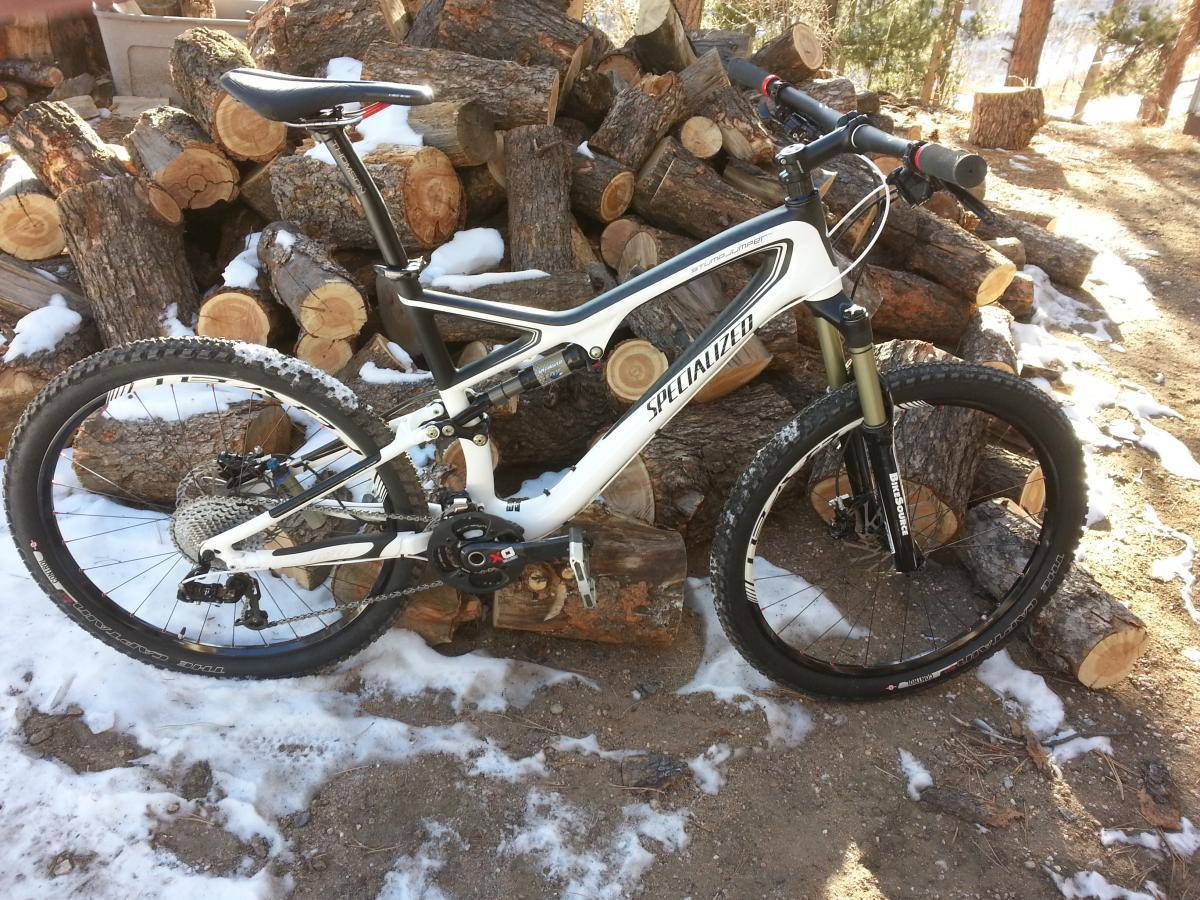 Specialized Stumpjumper FSR Pro Carbon: A white and black mountain bike leaning against a pile of cut logs, surrounded by snow and dirt. The bike features thick tires and a suspension system, situated in a natural outdoor setting with trees in the background.