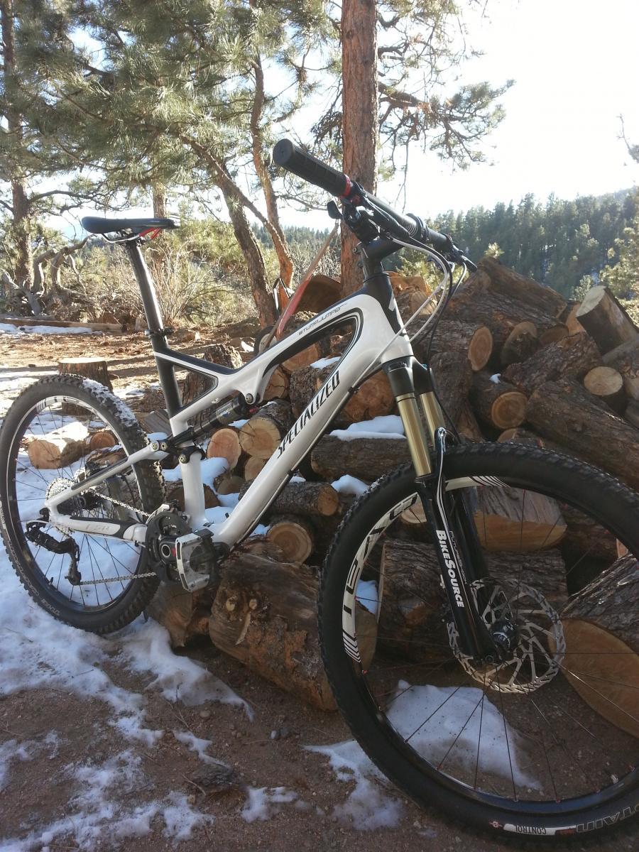 Specialized Stumpjumper FSR Pro Carbon: A white mountain bike is leaning against a pile of logs in a snowy outdoor setting, surrounded by tall trees and a clear blue sky.