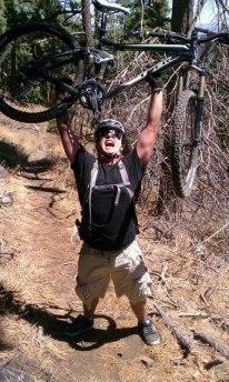 Trek Fuel EX 6: A person wearing a helmet and sunglasses triumphantly lifts a mountain bike above their head on a wooded trail, surrounded by trees and dry grass.