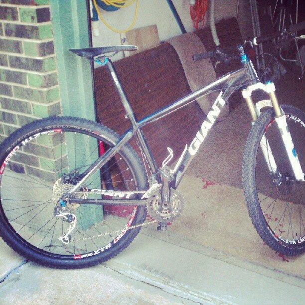 Giant XTC 1: A black and white mountain bike with the brand name "GIANT" prominently displayed on its frame. The bike features a front suspension fork and is positioned in a garage setting, with various tools and equipment visible in the background. The floor appears to be concrete, and there are some scattered items nearby.