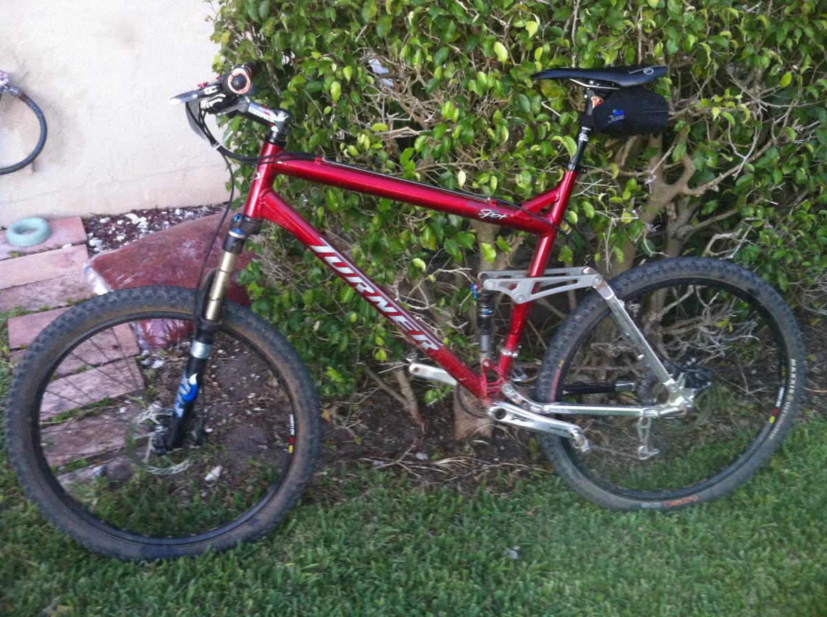 Turner 5.Spot: A red Turner Spy mountain bike leaning against a bush, with a partially visible tire in the foreground. The bike features a front suspension fork, disc brakes, and a small black pouch attached to the seat post. The background includes a patch of grass and some brick paving.