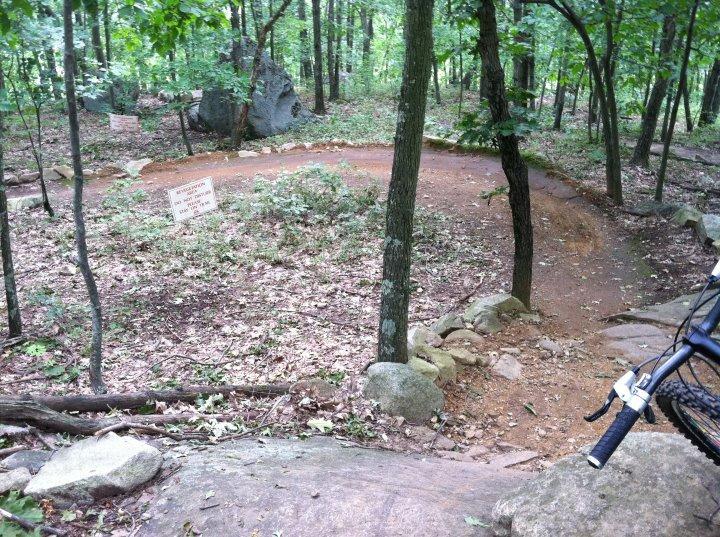 A winding dirt path surrounded by trees in a forested area, featuring signs indicating trail guidance. Rocks are scattered along the route, and a bicycle is partially visible in the foreground. Fallen leaves cover the ground, creating a natural, earthy atmosphere. Vietnam Trails mountain bike trail.