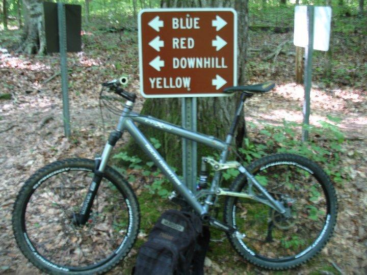A mountain bike leaning against a signpost in a wooded area, directing riders toward trails marked "Blue," "Red," "Downhill," and "Yellow." The ground is covered with leaves, and the scene is surrounded by greenery, indicating a natural outdoor setting. A black backpack is placed near the bike. Montgomery Bell State Park Mtb Trail mountain bike trail.