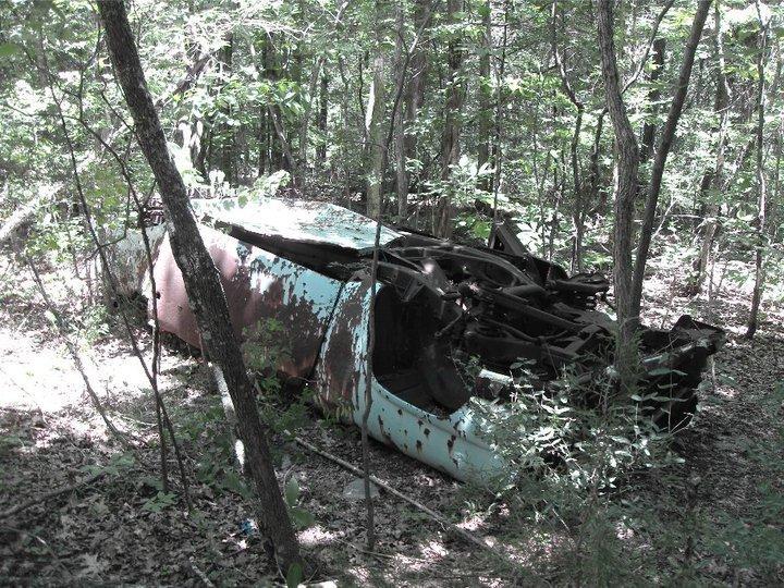 An abandoned, rusted vehicle partially hidden among dense foliage in a forest. The vehicle shows signs of decay, with its turquoise paint peeling and many rust spots visible. Sunlight filters through the trees, illuminating the scene. Hamilton Creek mountain bike trail.