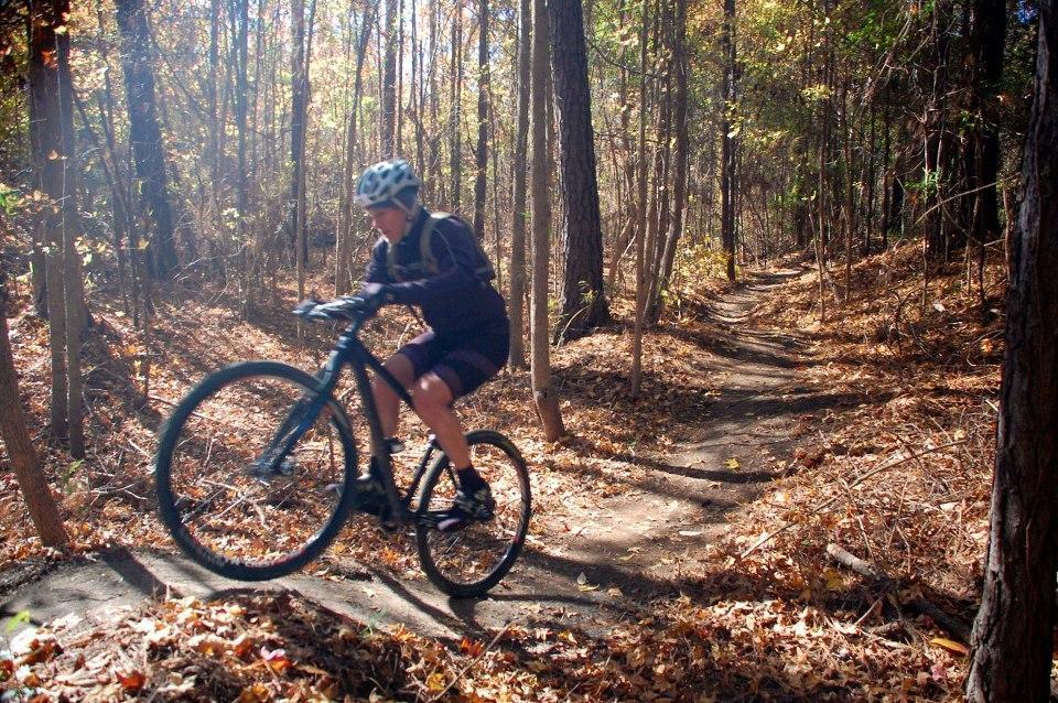 A cyclist in motion on a dirt trail, surrounded by trees and autumn foliage, with sunlight filtering through the leaves. The cyclist wears a helmet and is captured mid-jump on the bike. Horry County Bike Run Park mountain bike trail.