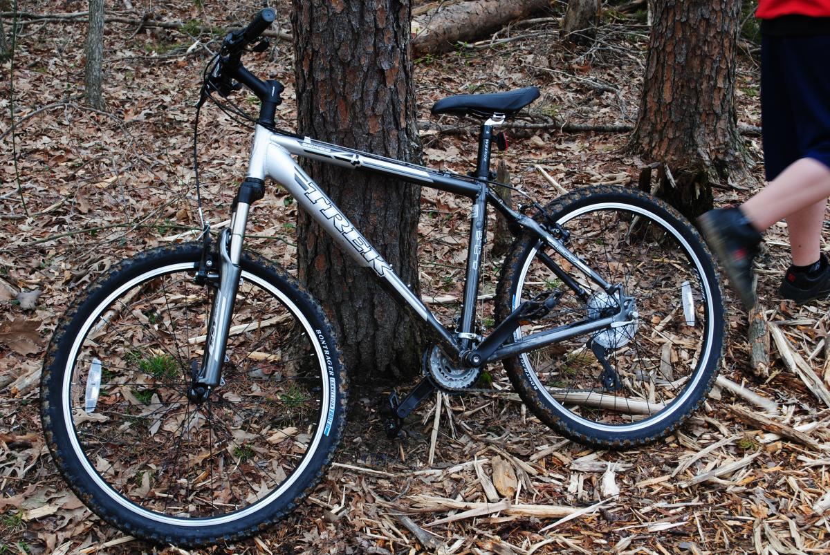 Trek 4300: A silver and black mountain bike leaning against a tree in a wooded area, surrounded by leaves and forest floor debris. A person in shorts and a shirt is partially visible in the background, walking past the bike.