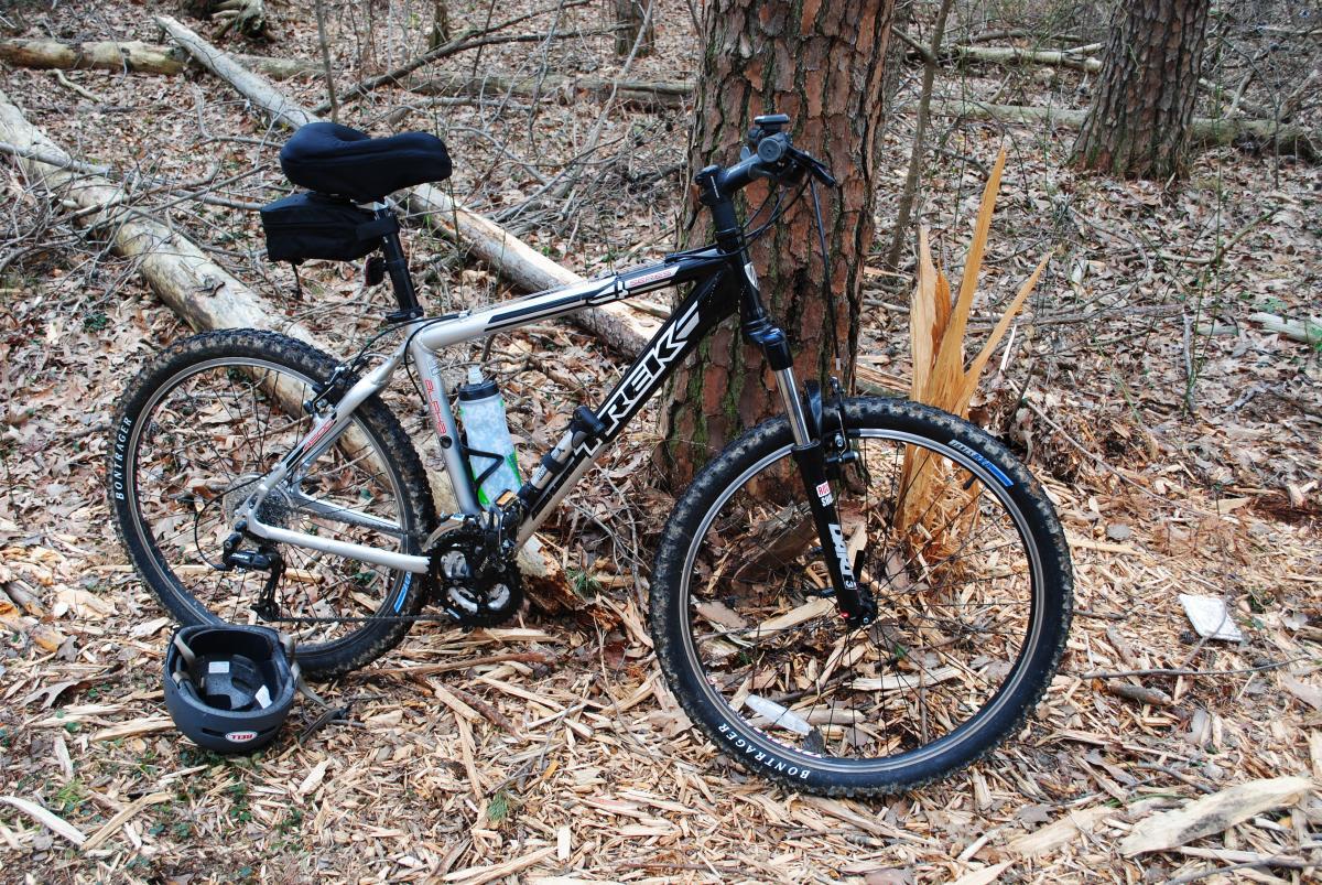 Trek 4500: A silver mountain bike leaning against a tree in a wooded area, with fallen leaves and branches in the background. A black helmet sits on the ground next to the bike, and a water bottle is mounted on the frame.