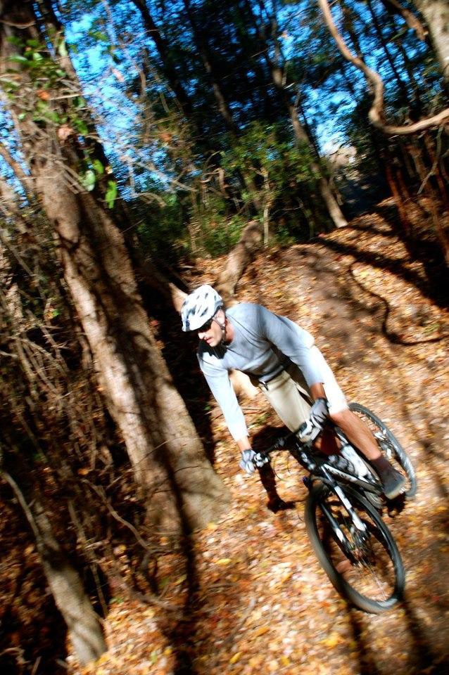 A cyclist in a light gray long-sleeve shirt and helmet rides swiftly along a wooded trail covered with autumn leaves, surrounded by tall trees and dappled sunlight. Horry County Bike Run Park mountain bike trail.