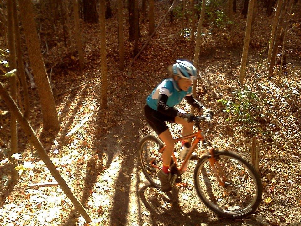 A child riding a mountain bike on a dirt trail surrounded by trees, with autumn leaves scattered on the ground and sunlight streaming through the branches. The child is wearing a blue cycling jersey and a helmet. Horry County Bike Run Park mountain bike trail.