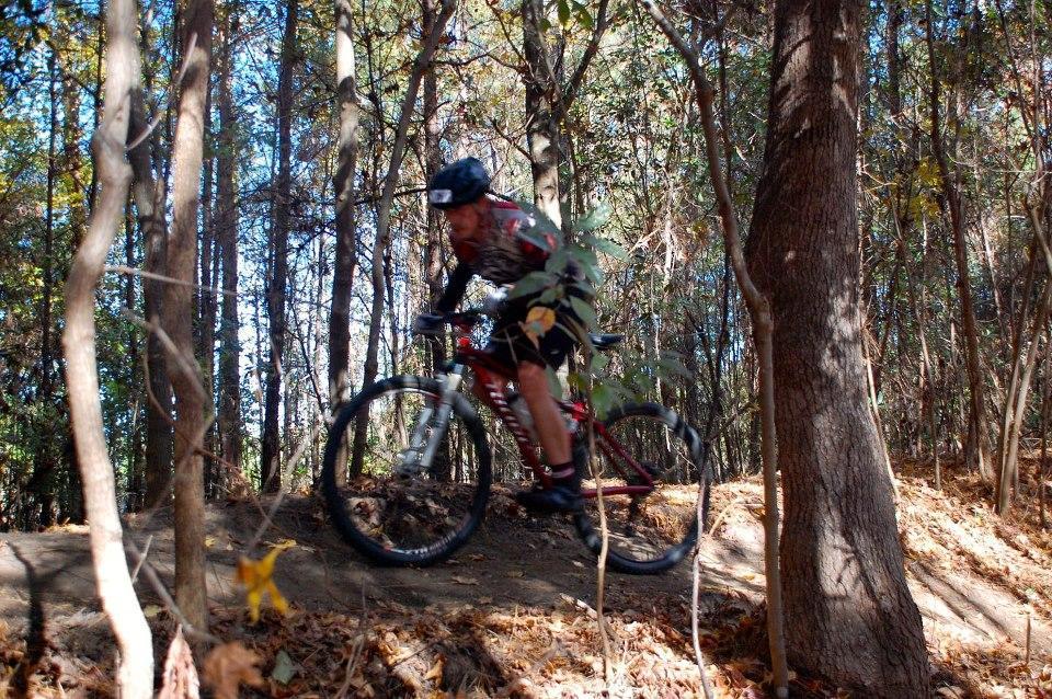 A mountain biker navigating a dirt trail surrounded by trees, with autumn leaves scattered on the ground. The cyclist is wearing a helmet and biking gear, captured in motion as they ride over a small incline in the forest. Horry County Bike Run Park mountain bike trail.
