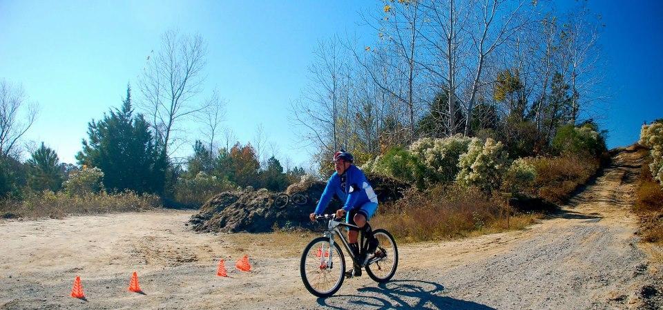A cyclist in a blue jacket and helmet rides a mountain bike on a dirt path surrounded by sparse trees and bushes. Orange cones mark the path, and a clear blue sky is visible above. The terrain appears rugged, with a slight incline ahead. Horry County Bike Run Park mountain bike trail.