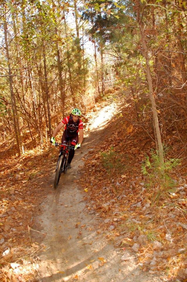 A mountain biker riding along a narrow dirt trail surrounded by trees in a forest, with autumn leaves covering the ground. The cyclist is wearing a red jersey, helmet, and gloves, navigating a slight incline. Horry County Bike Run Park mountain bike trail.