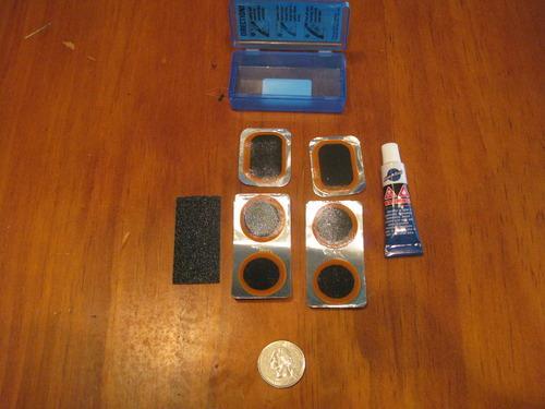 Park VP-1 Vulcanizing Patch Kit: A flat lay image featuring a small blue container with a hinged lid, a tube of adhesive, a gray sanding block, a quarter for scale, and four rectangular metal repair patches arranged on a wooden surface.