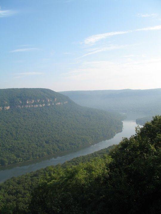 A scenic view of a river winding through lush green hills, with mist hovering over the landscape. The sky is clear with a few wispy clouds, creating a tranquil and serene atmosphere. The rugged cliffs on the left contrast with the gentle curves of the river below. Raccoon Mountain Trail Network mountain bike trail.