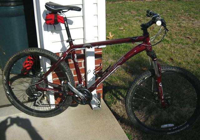 Gary Fisher Advance: A maroon mountain bike with thick tires, a suspension fork, and a visible gear system, parked on a concrete surface next to a brick wall and a grassy area. A red bag is attached to the bike's frame.