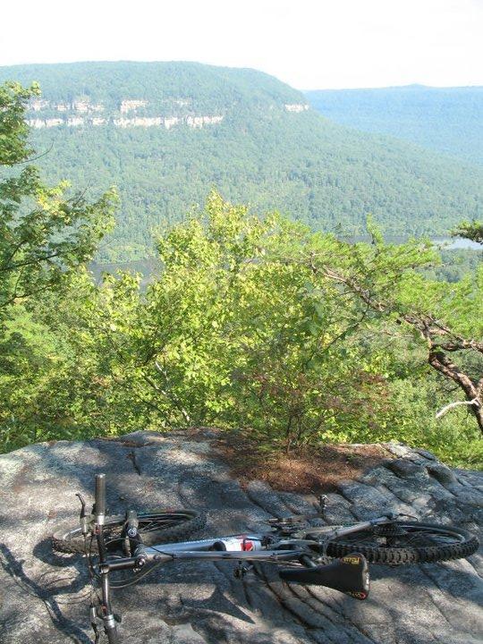 A mountain bike resting on rocky terrain, with a scenic view of lush green hills and cliffs in the background under a clear sky. Raccoon Mountain Trail Network mountain bike trail.