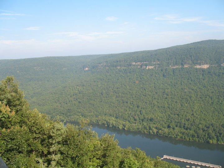 A panoramic view of a lush, green valley with rolling hills and a river winding through it. The scene is bathed in soft daylight, showcasing various shades of green in the trees, with blue skies and a few scattered clouds above. A small dock or structure can be seen along the river's edge, adding a focal point to the tranquil landscape. Raccoon Mountain Trail Network mountain bike trail.