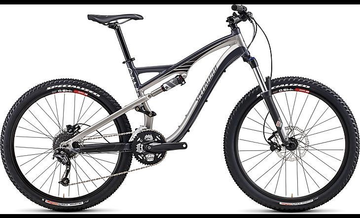 Specialized Camber: Image of a modern mountain bike featuring a dual suspension frame, sleek silver and black design, wide tires, and a gear system visible on the side. The bike is positioned upright against a plain background, showcasing its sturdy construction and performance-oriented design.