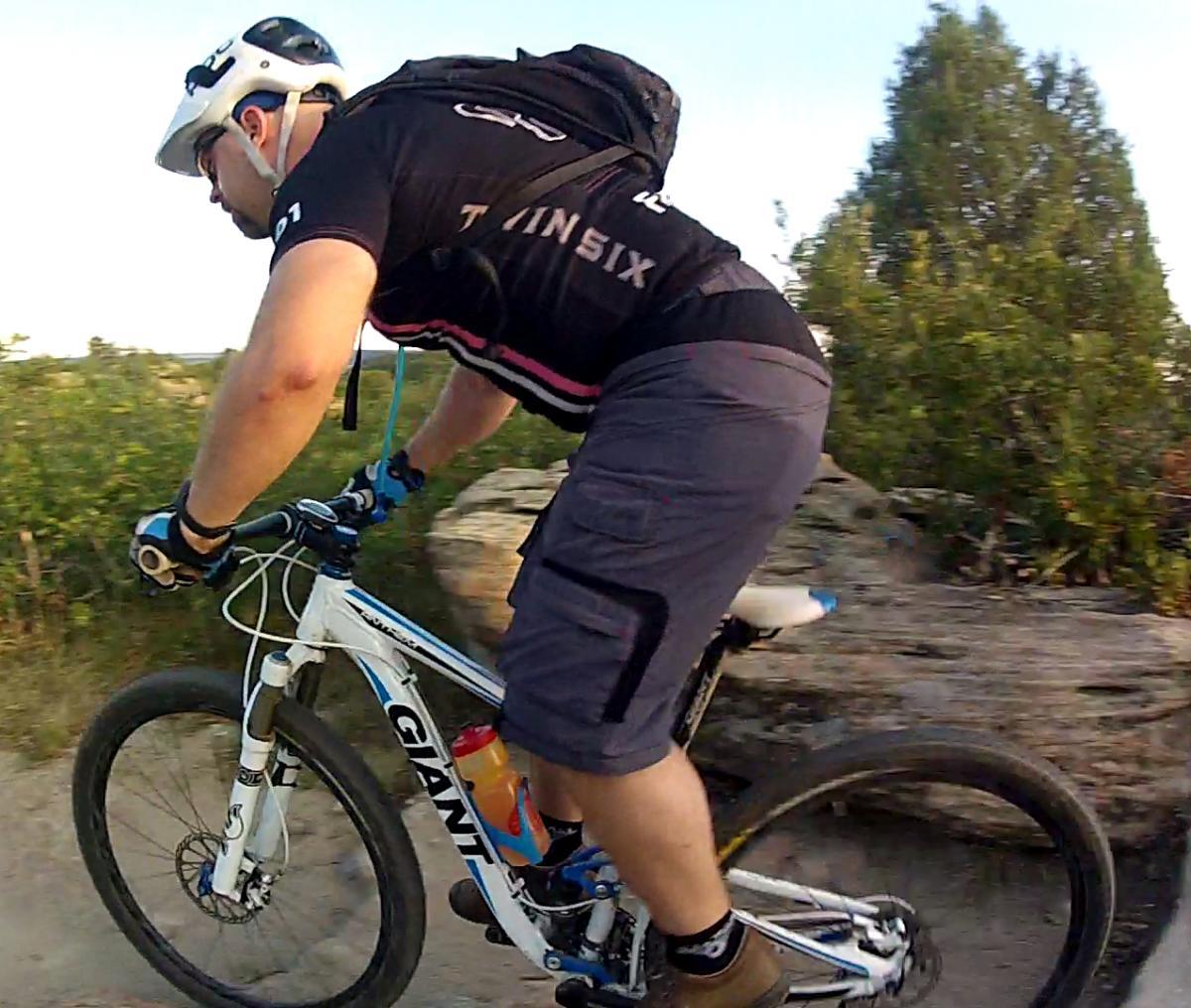 Giant Anthem X 29er 3: A cyclist riding a mountain bike on a rugged trail, wearing a helmet and athletic gear. The bike is white with branding visible, and the rider is navigating rocky terrain surrounded by greenery. The image captures the dynamic motion of cycling in an outdoor setting.