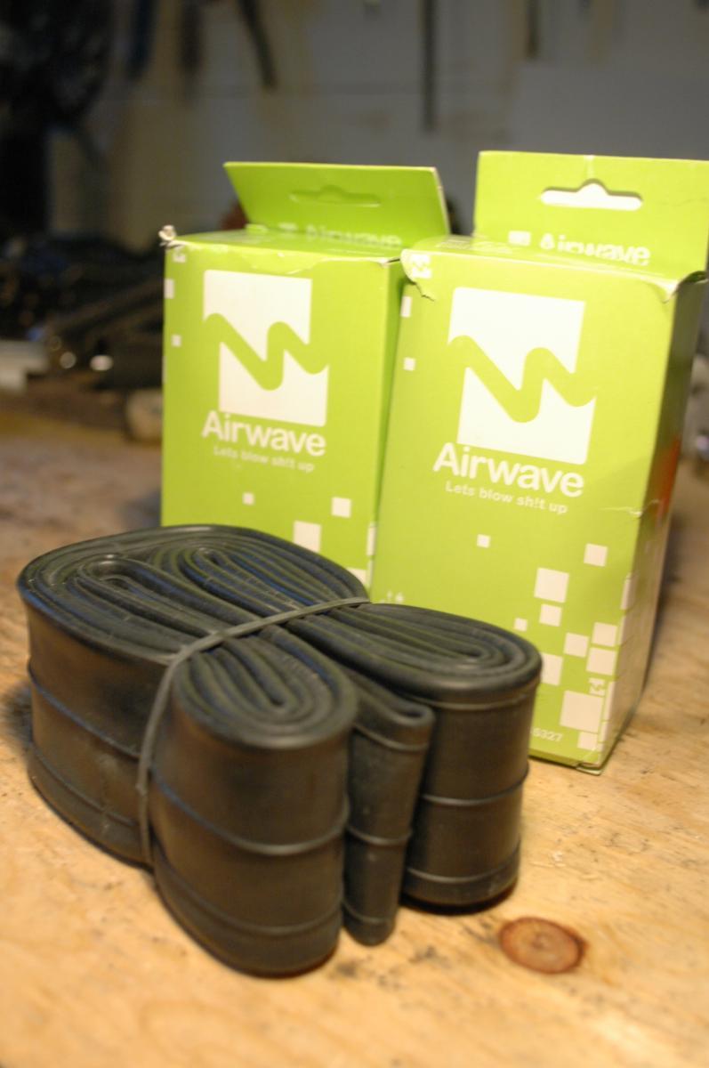 Airwave MTB Tube: Two green boxes labeled "Airwave" are visible in the background, accompanied by a bundle of black rubber tubes secured with rubber bands, placed on a wooden surface. The boxes communicate the slogan "Let