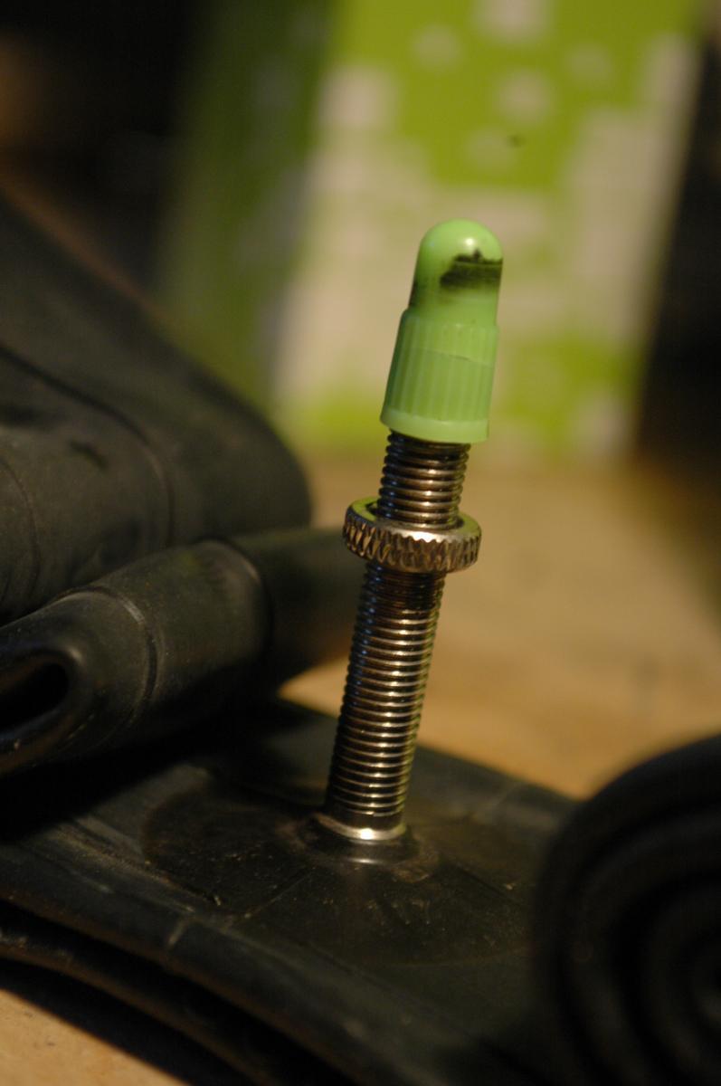 Airwave MTB Tube: Close-up of a bicycle valve stem mounted on a black inner tube, featuring a green cap at the top and surrounded by coiled rubber. The background is blurred, highlighting the valve assembly.