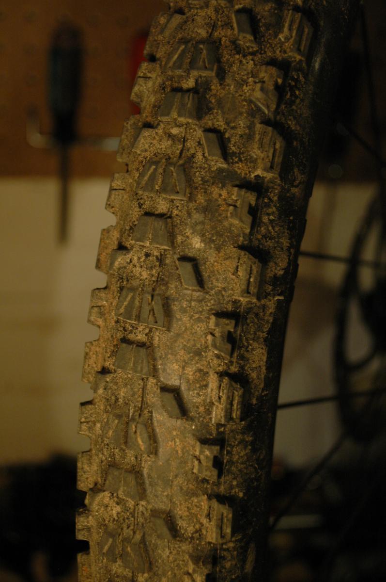 Maxxis Ardent: Close-up of a muddy mountain bike tire with a rugged tread pattern, set against a blurred background in a garage environment.