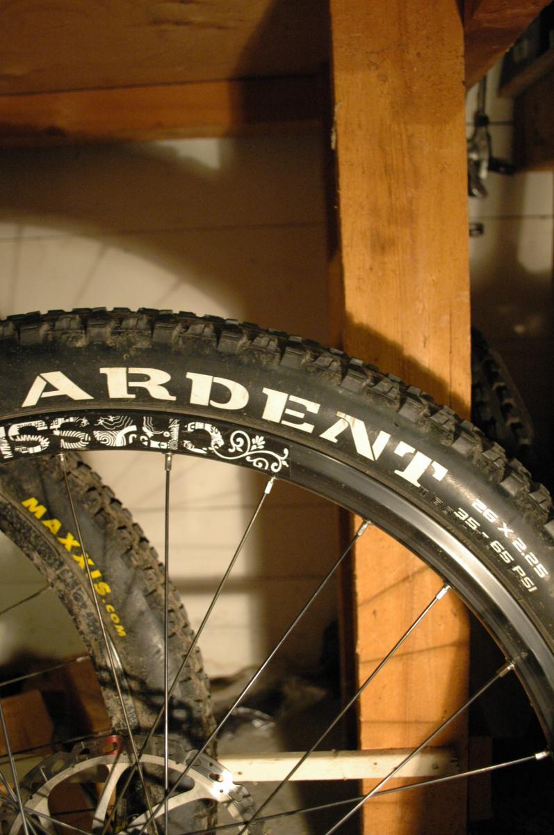 Maxxis Ardent: A close-up image of a mountain bike tire labeled "Ardent" mounted on a wheel, with a visible tire tread pattern. The tire specifications reading "26 x 2.25" and recommended pressure are also displayed. In the background, another tire with a yellow "Maxxis" logo is partially visible, along with a wooden shelf and tools in a garage setting.