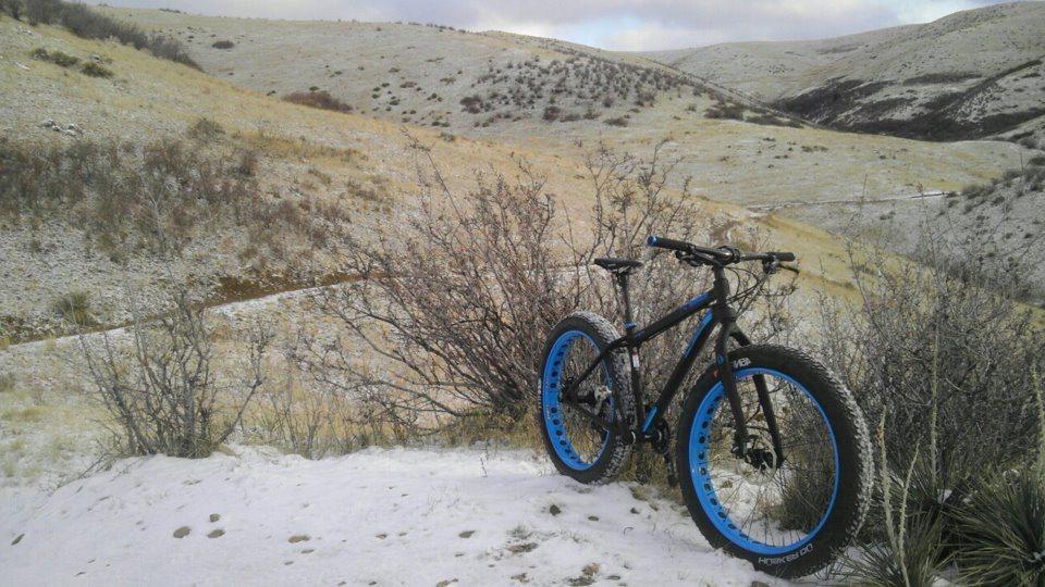 A fat tire bike with blue rims resting on a snowy patch of ground, surrounded by shrubs and rolling hills in a winter landscape. The sky is overcast, suggesting a cold day in a remote outdoor area. Green Mountain mountain bike trail.