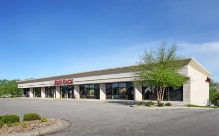 Exterior view of a commercial building with large glass windows and a flat roof, featuring a sign that reads "Rents & More." Surrounded by green landscaping and a clear blue sky.