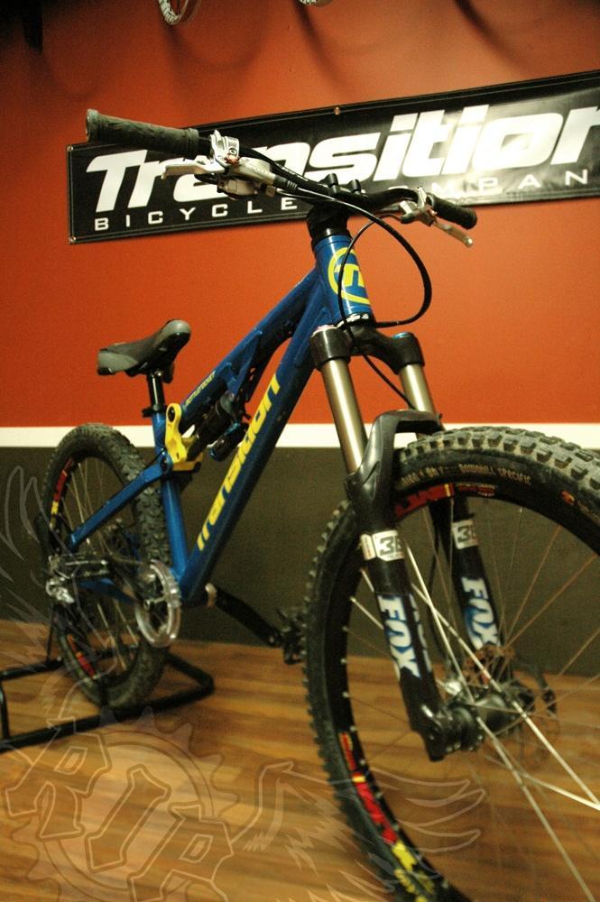 Transition Bottlerocket: A blue transition mountain bike displayed in a shop setting, featuring a sturdy frame, wide tires, and front suspension, with a 'Transition Bicycle Company' sign in the background and wooden flooring.